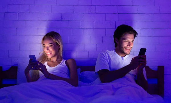 Fiatal házastársak használata telefonok fekvő ágyban Night — Stock Fotó