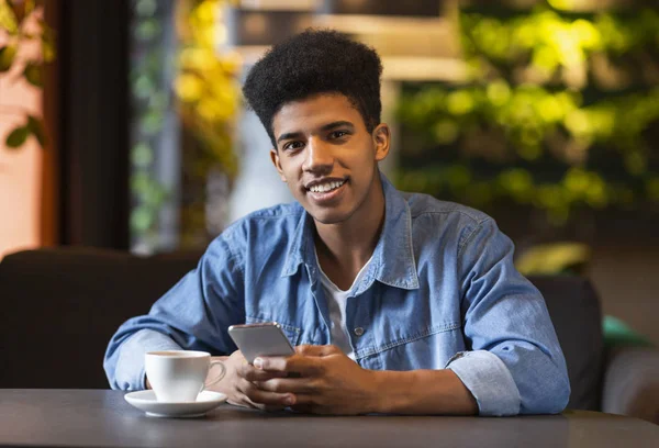 Przystojny czarny nastolatek facet trzyma telefon komórkowy, uśmiechając się w aparacie — Zdjęcie stockowe