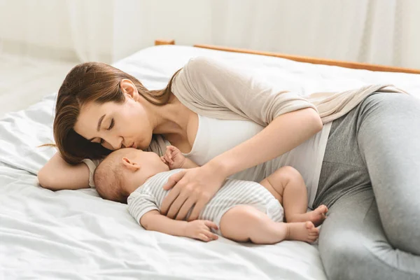 Young mother kissing her sleeping newborn baby in bed. — Stock Photo, Image