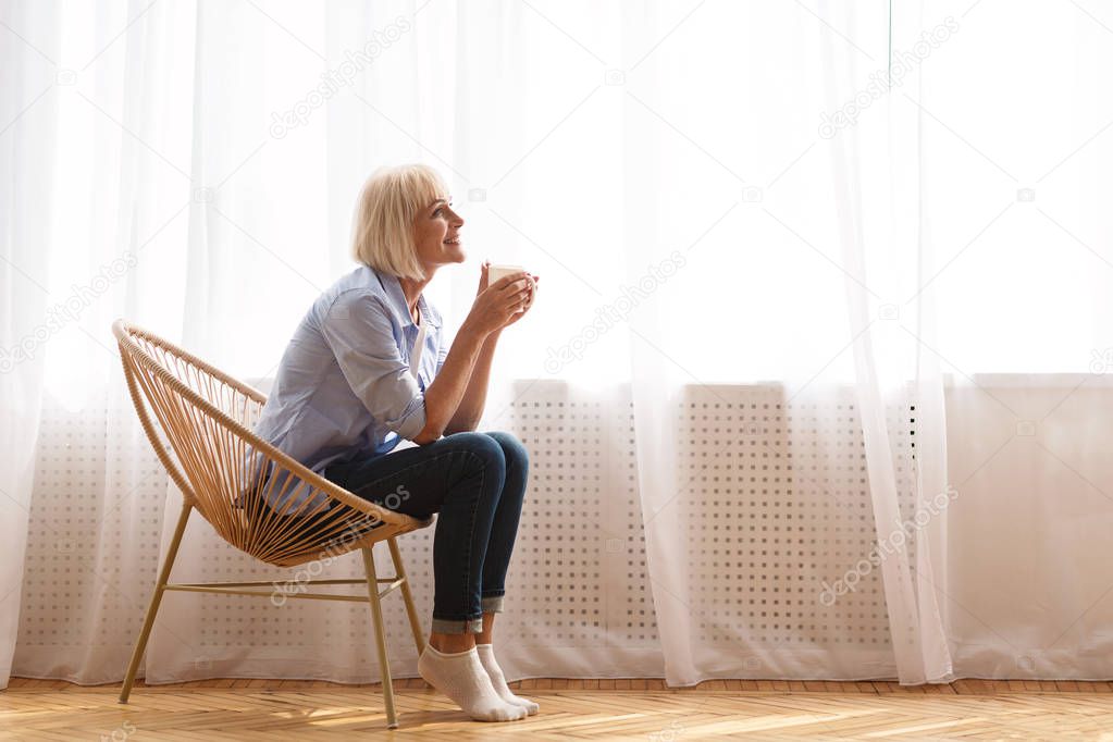 Senior woman sitting on chair in front of window and drinking tea