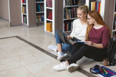 Kütüphanede yerde oturan iki kız arkadaş, dizüstü bilgisayarla çalışıyor.