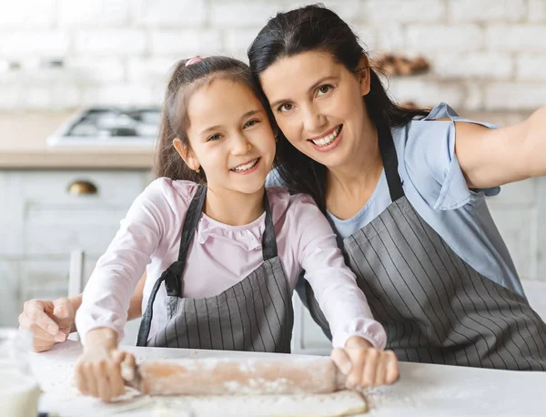 Niedliche kleine Mädchen und ihre schöne Mutter in Schürzen Selfie machen, lächelnd in der Küche — Stockfoto