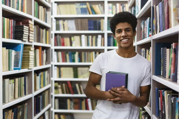 Alegre afro-americano cara ficar entre estantes, segurando livro — Fotografia de Stock