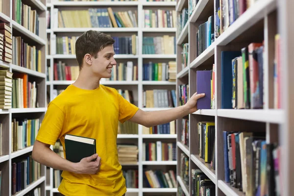 Ung kaukasiska kille som tar bok från bokhyllan — Stockfoto