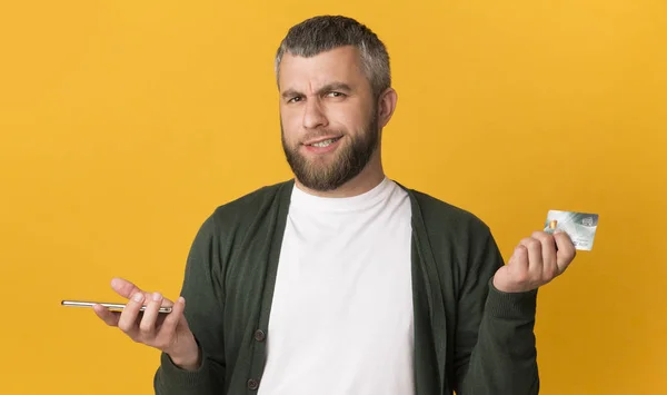 Hombre escéptico sosteniendo teléfono celular y tarjeta de crédito —  Fotos de Stock