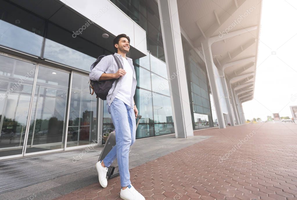 Happy millennial man walking with luggage out of airport building