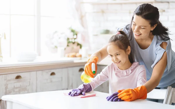 Caring mom teaching daughter how to clean table with detergent