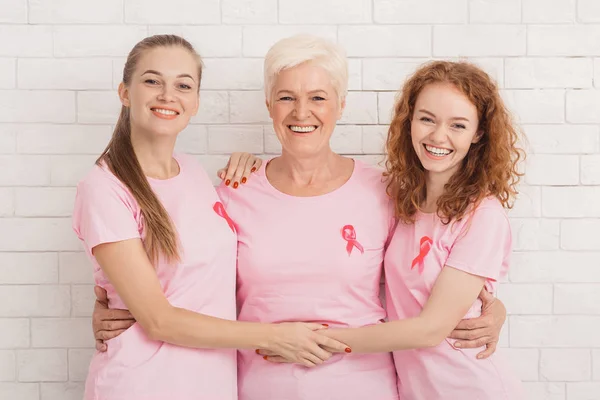 Tres mujeres con camisetas de cáncer de mama abrazándose contra la pared blanca —  Fotos de Stock