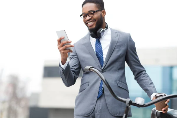 Empreendedor afro feliz usando telefone, de pé com bicicleta ao ar livre — Fotografia de Stock
