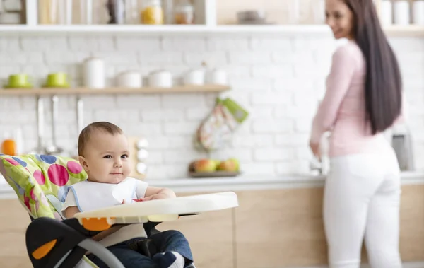 Schattige baby spelen op kinderstoel in keuken, moeder wassen gerechten — Stockfoto