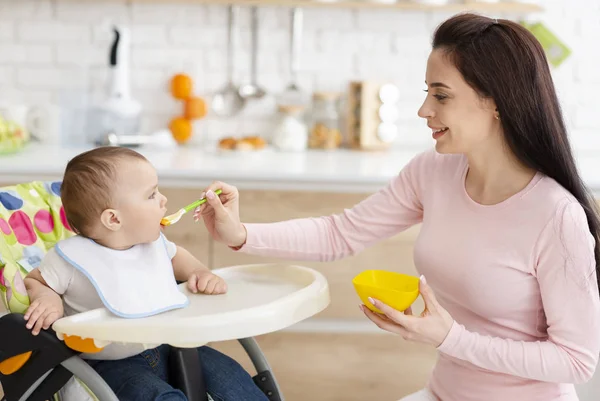 Mãe alimentando seu bebê, criança sentada em cadeira alta na cozinha — Fotografia de Stock