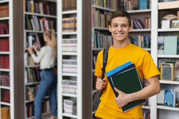 Захоплений студент позує поруч з книжковими полицями в університетській бібліотеці — стокове фото