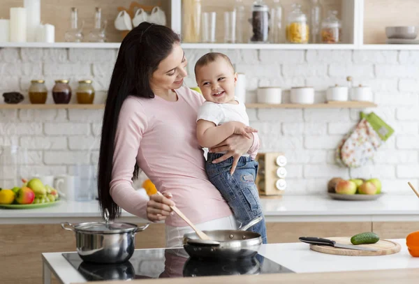 Donna millenaria che ride con figlio neonato, prepara il cibo in cucina — Foto Stock
