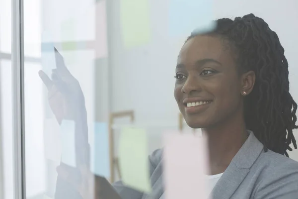 Afro signora mettendo appiccicoso note su vetro bordo a luogo di lavoro — Foto Stock