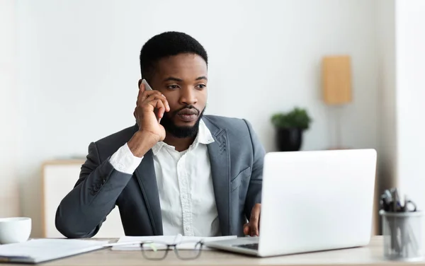 Koncentruje Afro biznesmen mówić na telefon komórkowy i pracy na laptopie — Zdjęcie stockowe