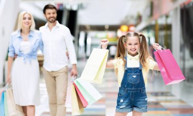 Alışveriş merkezinde yürüyen alışveriş poşetleri taşıyan ebeveynler ve heyecanlı kızı
