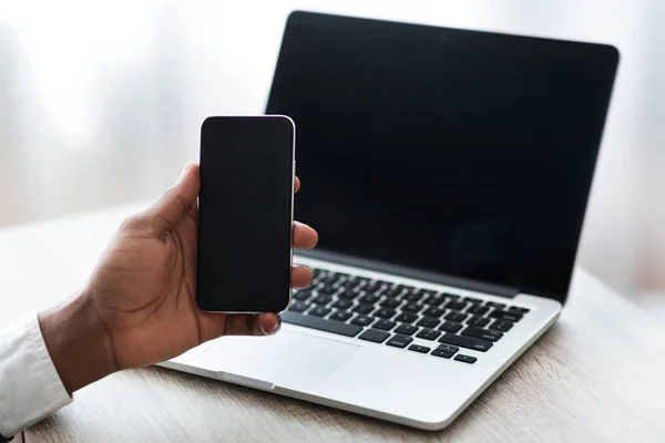 Чорний чоловік тримає смартфон з порожнім екраном, використовуючи ноутбук — стокове фото