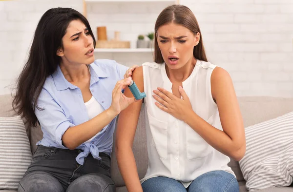 Girl Giving Asthma Inhaler To Friend Sitting On Couch At Home — Stock Photo, Image