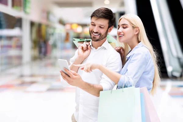 Coppia sorridente che utilizza il cellulare alla ricerca di offerte di vendita in piedi nel centro commerciale — Foto Stock