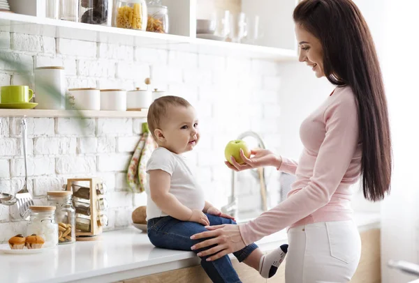 Caring mother offering fresh green apple to her infant at kitchen — Stock Photo, Image