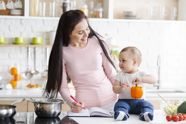 Mama notiert Gedanken, sitzt mit Baby in der Küche — Stockfoto