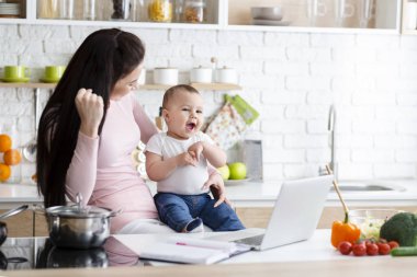 Mutlu kadın başarı zevk, mutfakta bebek ile dizüstü bilgisayar üzerinde çalışıyor