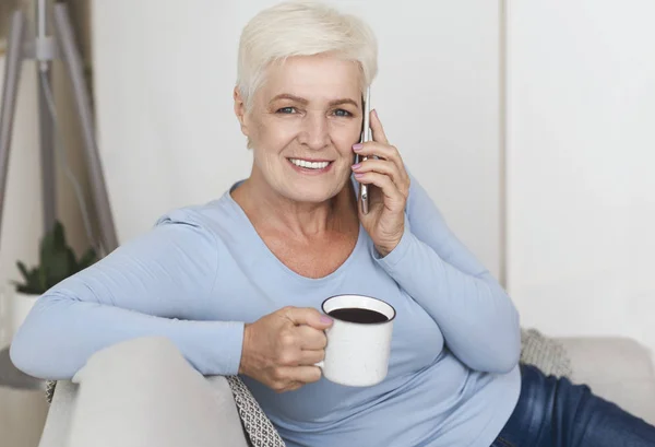 Attraktive alte Frau trinkt Kaffee und telefoniert mit dem Smartphone — Stockfoto