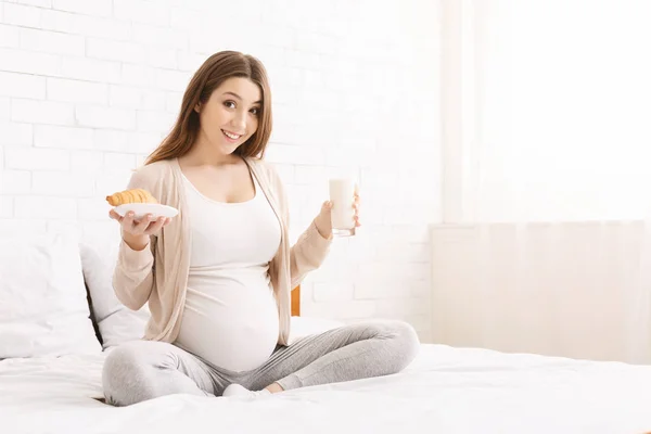 Pregnant woman enjoying milk and croissant, sitting on bed — Stock Photo, Image