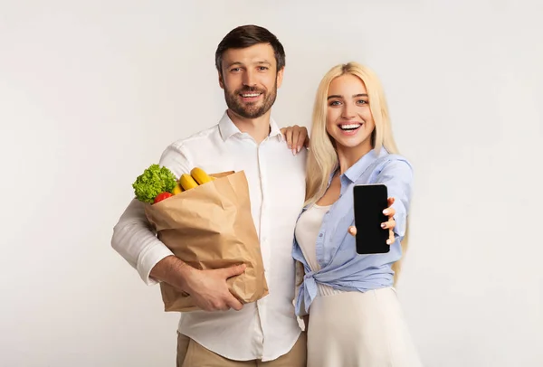 Casal mostrando tela do celular segurando o saco de compras do supermercado, tiro do estúdio — Fotografia de Stock