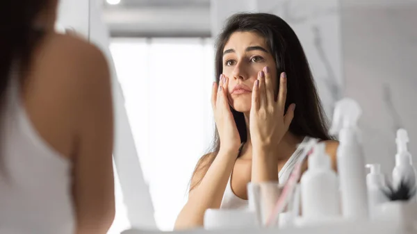 Женщина касается лица, глядя на кожу в зеркале в ванной комнате — стоковое фото