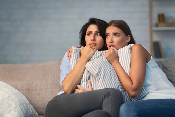 Due ragazze spaventate abbracciano cuscini seduti sul divano a casa — Foto Stock