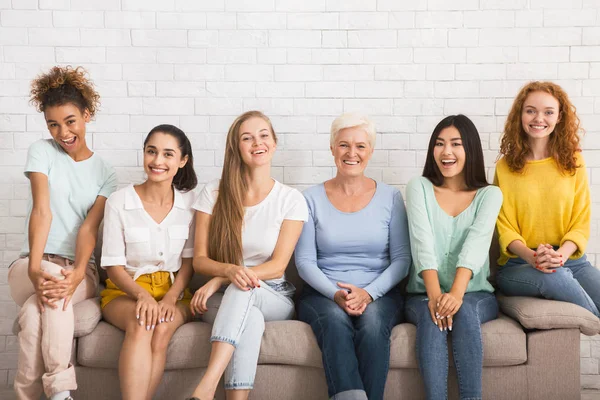 Uśmiechnięty różnorodny kobiety siedzący na sofa przez biały cegła ściana — Zdjęcie stockowe
