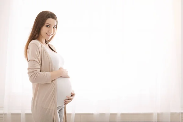 Fiatal terhes nő ölelgeti a hasát, az ablak mellett áll — Stock Fotó