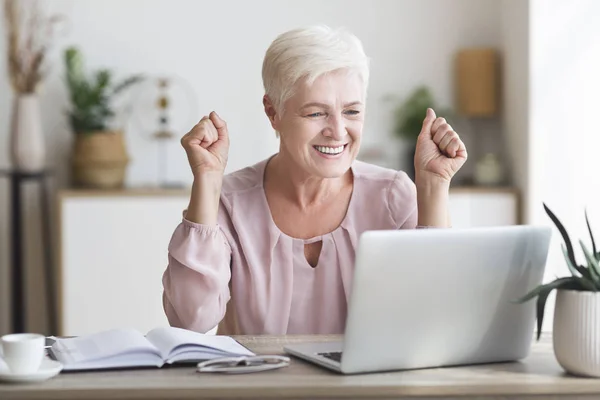 Щаслива старша жінка святкує успіх перед ноутбуком — стокове фото