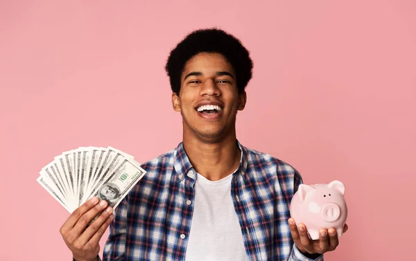 Siyah adam pembe arka plan üzerinde para ve kumbara tutan — Stok fotoğraf