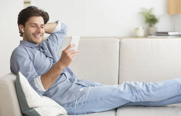 Man Using Smartphone Listening To Music Lying On Sofa Indoor