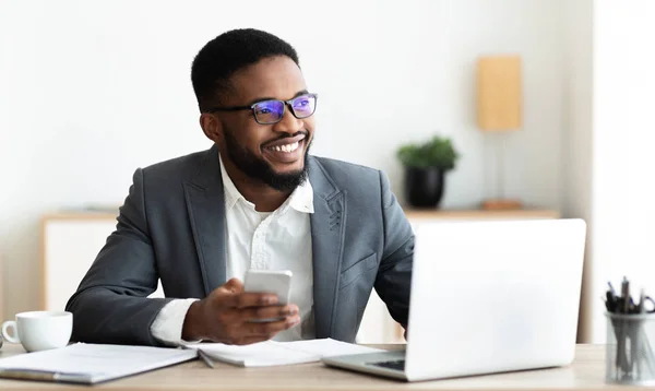 Portret uśmiechu czarny biznesmen siedzi w miejscu pracy z telefonem komórkowym — Zdjęcie stockowe