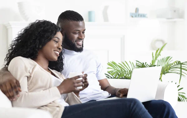 Cheerful black couple sitting on couch looking at laptop screen — ストック写真