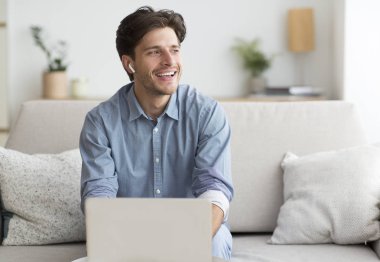 Kulaklık takan, dizüstü bilgisayar kullanan evdeki koltukta oturan adam.