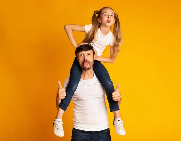 Zabawny ojciec niosący córkę na ramionach Pokazywanie języków, Żółte tło — Zdjęcie stockowe