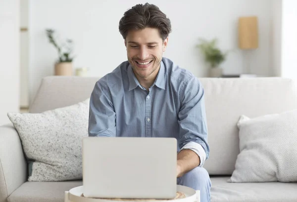 Hombre sonriente trabajando en el ordenador portátil sentado en el sofá en casa — Foto de Stock