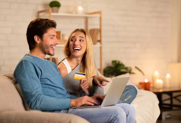 Casal segurando laptop e cartão de crédito sentado no sofá interior — Fotografia de Stock