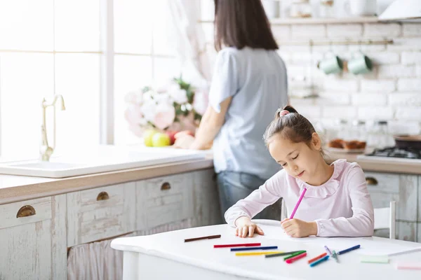 Schattig meisje tekening aan keukentafel terwijl haar moeder koken — Stockfoto