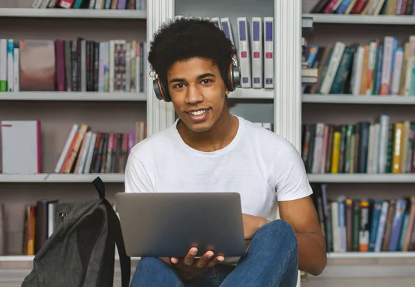 Étudiant afro-américain avec casque appuyé sur la bibliothèque, à l'aide d'un ordinateur portable — Photo