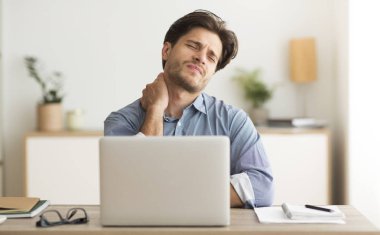 Boyun ağrısı çeken genç adam iş yerinde dizüstü bilgisayarda çalışıyor.