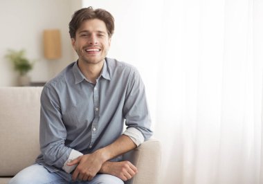 Genç adam kameraya gülümsüyor Evdeki koltukta oturuyor