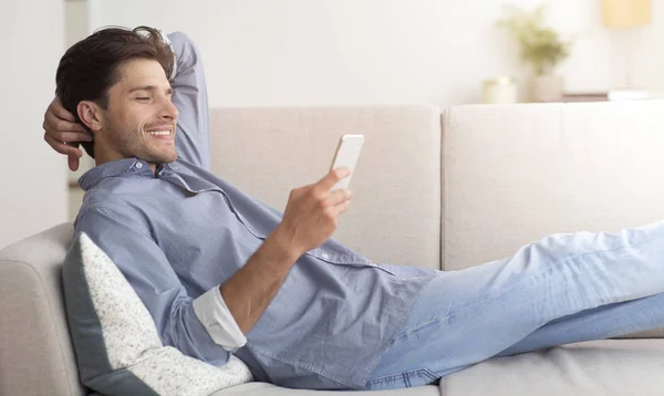 Homem relaxado navegando no smartphone deitado no sofá em casa — Fotografia de Stock