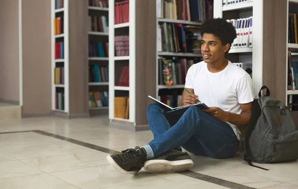 Kreatywny afro student robi notatki, siedzi na podłodze w bibliotece — Zdjęcie stockowe