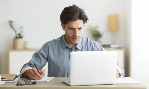 Joven emprendedor trabajando en el ordenador portátil y tomando notas en interiores — Foto de Stock