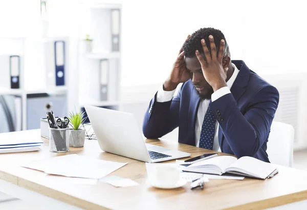 ストレスを受けたアフリカのビジネスマンは、額に手をつないでオフィスに座っている — ストック写真